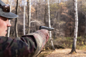 A man in camouflage clothes aims from a gas pistol in his hands in the forest in autumn