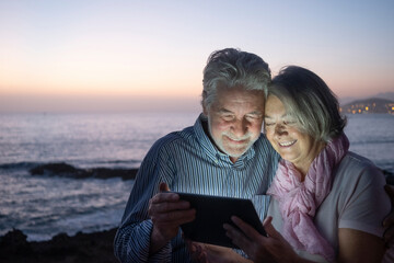 Cheerful senior couple white-haired in video chat with a digital tablet, sitting in the dusk on the beach with horizon over water on background. Happy retirement concept together