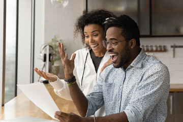Happy young African American couple feel euphoric read good unbelievable news in paper letter...