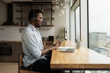 Young biracial man in headphones talk on video call on laptop in home office, make notes in notebook. African American male employee in earphones have webcam digital virtual conference online.