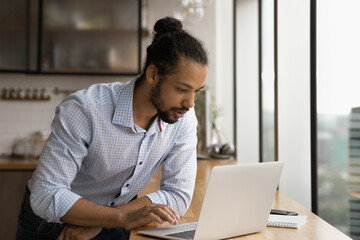 Young African American man look at laptop screen working online in office. Concentrated millennial...