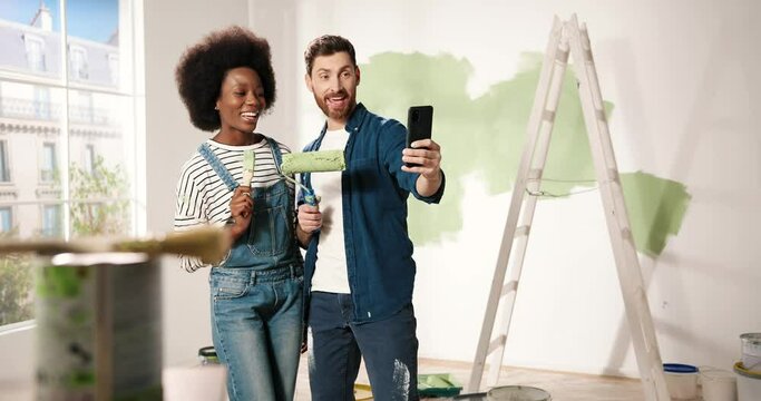 Joyful mixed-races young family couple wife and husband smiling posing with brushes taking selfie photos on smartphone in room. Home repair concept. Man and woman taking pictures during renovation