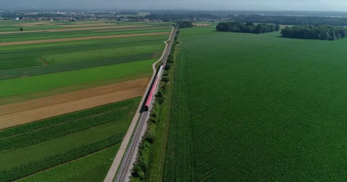 Aerial drone flying over large farming grassland fields, Slovenia. Elevated view of flatland farmland. Passenger train passing by on straight railway railroad between agriculture fields. Forward dolly