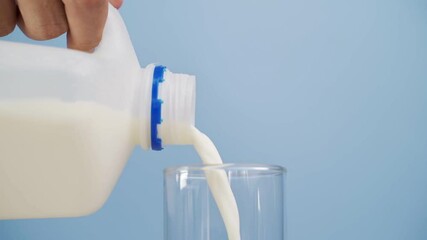close up hand pouring milk from gallon into glass on blue background.