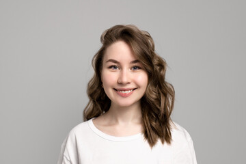 Portrait of happy smiling young woman looking at camera with natural everyday makeup, gray background, copy space. Beautiful caucasian girl with brown curly hairstyle. Cheerful expression face