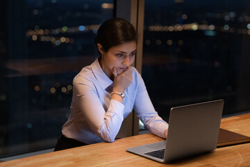 Pensive young Indian woman sit at desk look at laptop screen work in office late hours alone....