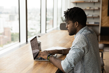 Serious millennial African American man sit at desk at home office look at laptop screen working...