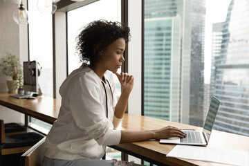 Pensive millennial biracial woman sit in modern office work on modern laptop thinking. Thoughtful...