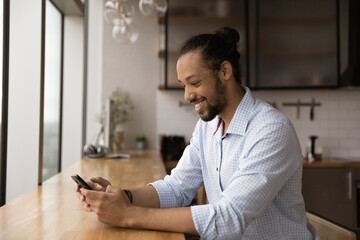 Obraz na płótnie Canvas Smiling millennial African American man sit at desk in office or home look at cellphone screen browsing internet. Happy young biracial male use smartphone texting or messaging on gadget online.