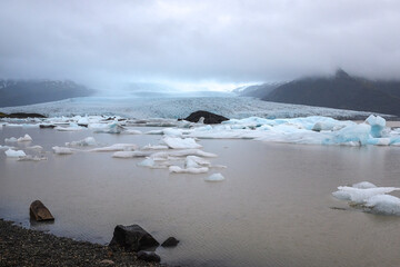 view of the glacier from the shore of the lake on a rainy day with an iceberg floating on the lake