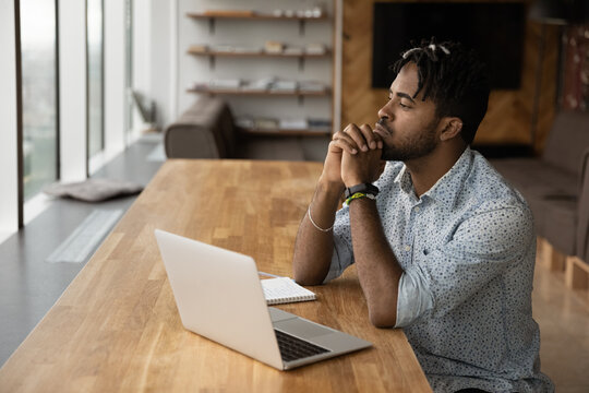 Thoughtful young African American man sit at desk in home office distracted from computer work pondering. Pensive biracial male use laptop thinking or planning, solve problem or make decision.