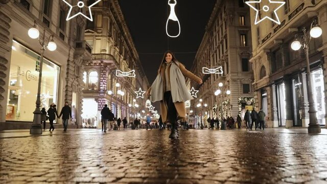 Millennial girl in a beige long coat and scarf walks along the evening street in the winter Milan city. Woman wearing warm clothing