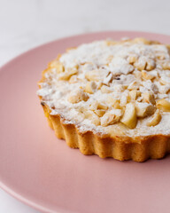 Delicious apple tartlet with caramelized apple, custard cream, roasted nuts and powdered sugar served on a pink plate with light background. Selective focus. Copy space.