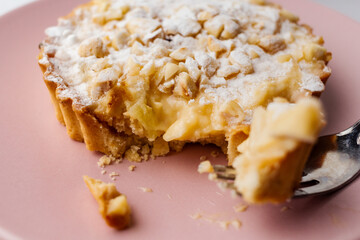 Delicious apple tartlet with caramelized apple, custard cream, roasted nuts and powdered sugar served on a pink plate with light background. Selective focus. Copy space.