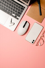 Vertical flat lay shot with a grey laptop, notebook, pen, white mouse, modern smartphone, stylish goggles, and wireless earphones on a pink background.