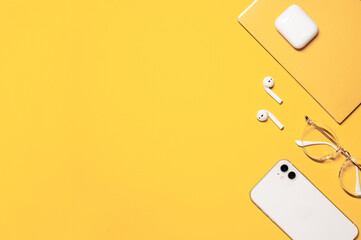 Flat lay photo with a copy space on the left side, and objects on the right: white smartphone, wireless earphones with a charging case, stylish goggles, and a yellow notebook on a yellow background.