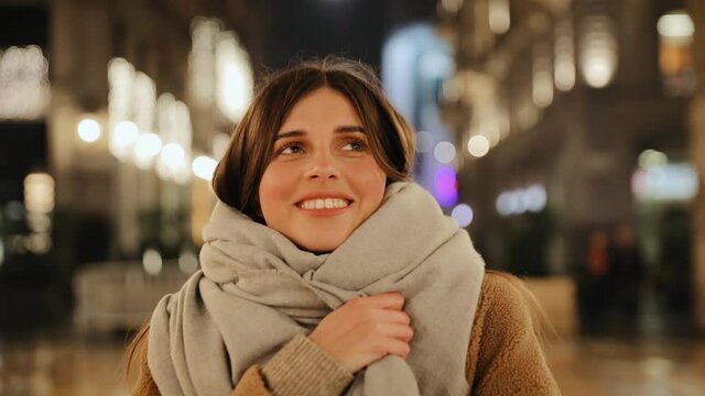 Pretty young woman in a coat and with a scarf is walking in the night city. Walk along the evening streets in the autumn. Portrait view. She is close her eyes and smile during walking