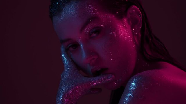 Sensual girl in sparkles in neon light posing in studio, sexy female with glittering make up and bare shoulder