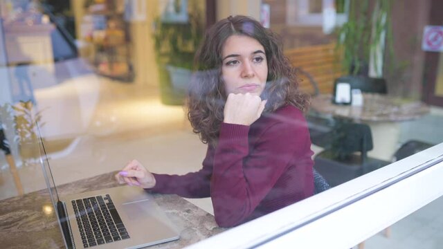 Young caucasian woman portrait sitting in a cafe with computer laptop looking out the window with reflection on glass with thoughtful face. Technology, business, remote working or studying concept.