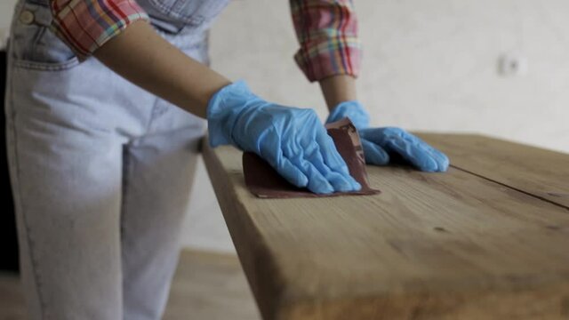 Unrecognizable female woodworker in gloves polishing lumber table with sandpaper while working in local carpentry workshop 