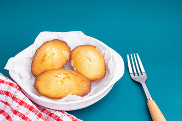 Lemon glaze madeleines on a plate placed on a blue light background. Madeleine - homemade traditional French small cookie. Seashell sweet cakes