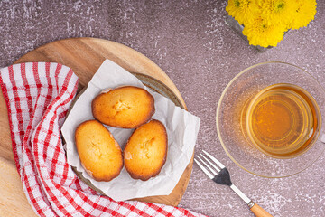 Lemon glaze madeleines on black a plate with a cup of tea placed on a marble background with a white brick wall. Madeleine - homemade traditional French small cookie. Seashell sweet cakes