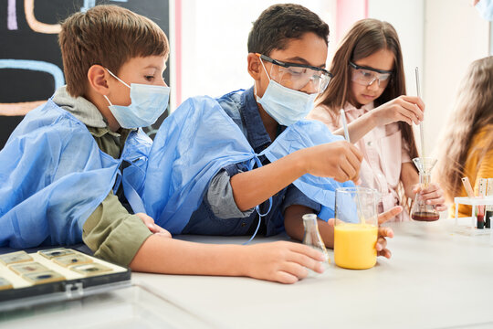 Two Curious Boys In Protective Masks And Gowns Mixing Liquids