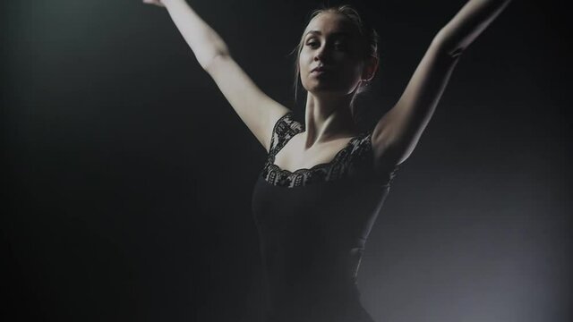 Young woman ballerina in black dress slowly turning around on the smoky stage