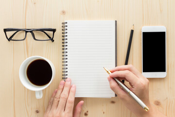 Female hand writing on notebook surrounded with office accessories. Top view of work desk. Coffee, pencil, paper