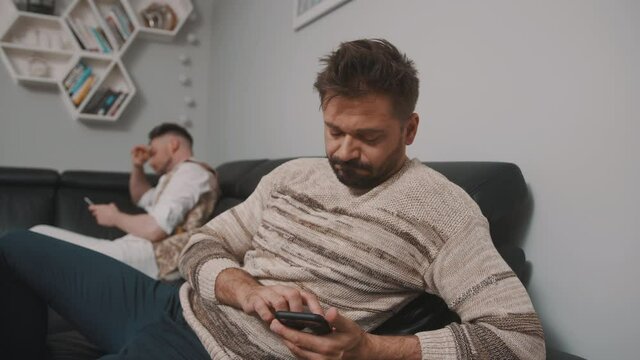 Gay couple sitting on the oposite sides of the couch and looking at their phones. Difficult times in relationship. High quality 4k footage