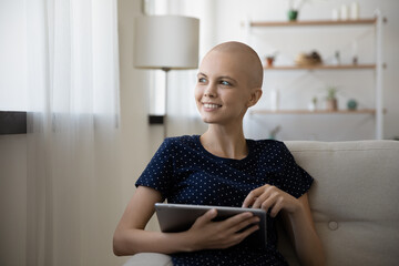 Optimistic glad young female cancer patient rest on couch with digital tablet distracted from...