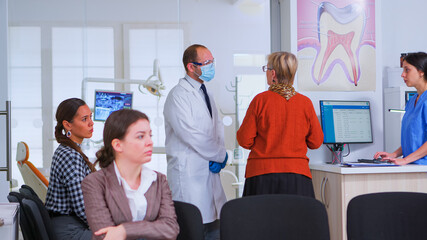 Dentistry doctor talking with senior woman standing in reception area while patients waiting sitting on chairs in crowded orthodontist clinic. Dental nurse typing on computer making appointments.