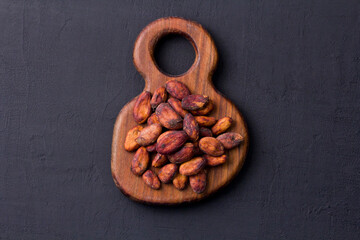 Unpeeled raw brown cocoa beans in heap lie on wooden board, on black background. Flat lay, mock up, copy space. Raw materials for making cocoa powder, cocoa beverages, chocolate. Health drink concept