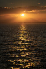 The sun doing down over a darkening ocean, making the waves sparkle. Distant land is visible on the horizon