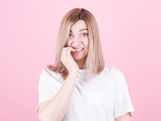 Close up portrait of a worried teenage girl in white t-shirt biting her nails isolated over pink background