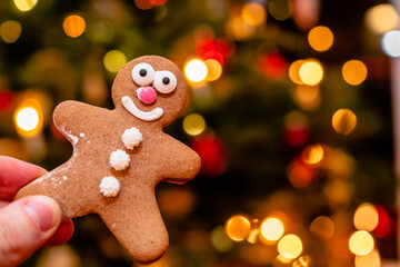 Obraz na płótnie Canvas Hand holding a smiling gingerbread man in front of a blurred background with warm bokeh lights. Christmas lights background. Celebrating winter holidays, advent, Christmas season. Festive decoration.