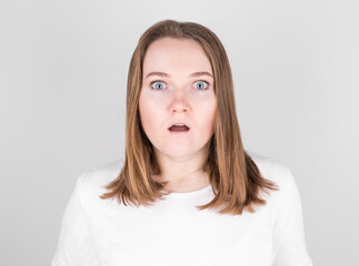 Young woman in a white T-shirt stands against a gray wall in great surprise.