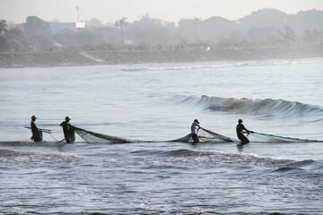 Group of balinese fishermen pulling large fishing net together on sea shore.