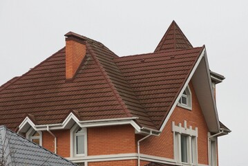 brick house with white windows and brown tiled roof with chimneys against gray sky