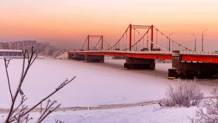 Fotobehang Purper A soothing winter landscape at sunset with a bridge over a frozen river against a backdrop of city buildings and an orange sky.  © Aleksandr 44ARH