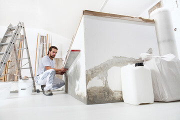 construction worker plasterer man looks at the spirit level and checks the wall in building site of home renovation with tools and building materials on the floor