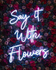 Say with the flowers sign. Neon sign on the wall. Motivation quotes background. Neon words sign. Flowers and gift concept. Background with fresh flowers. 