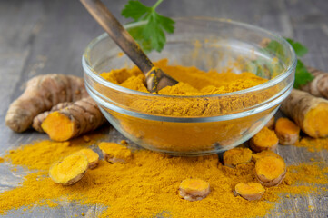 Fresh turmeric root and turmeric powder in a transparent bowl on a gray background.