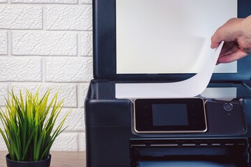 A young man's hand lays a stack of white paper on the scanning surface of a black printer. Green...