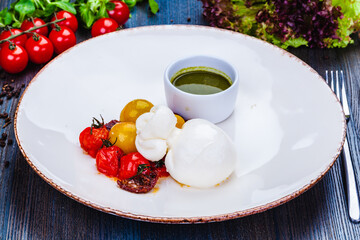 Burrata cheese with baked cherry tomatoes and pesto sauce on white plate.