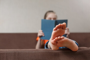 A funny little caucasian girl with bare feet with book while sitting on a sofa at home. Foot...