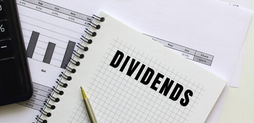 Text DIVIDENDS on the page of a notepad lying on financial charts on the office desk.