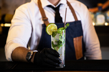 Detail of the hands of a bartender with black gloves placing a crystal glass with a cocktail inside, on a bar counter