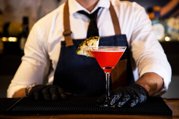 Detail of the hands of a bartender with black gloves placing a crystal glass with a cocktail inside, on a bar counter