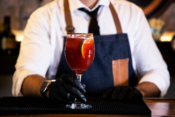 Detail of the hands of a bartender with black gloves placing a crystal glass with a cocktail inside, on a bar counter